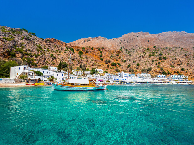 A wooden boat floats on clear turquoise water near a coastal village with white buildings, set against arid, rocky mountains and a vibrant blue sky.