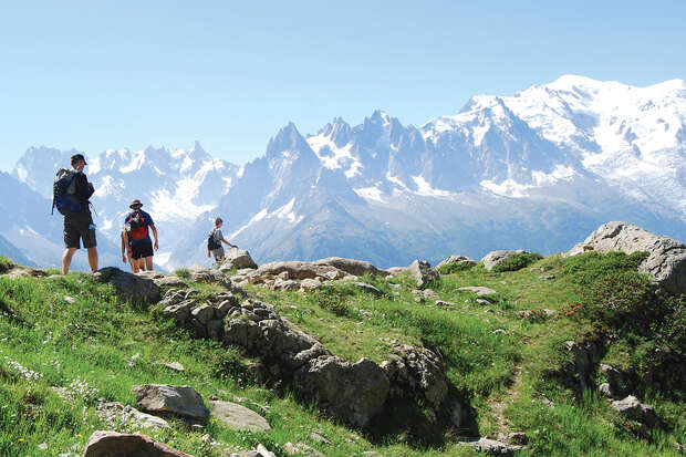 Hikers pause on a lush, rocky hillside, overlooking a majestic range of snow-capped mountains under a clear blue sky.