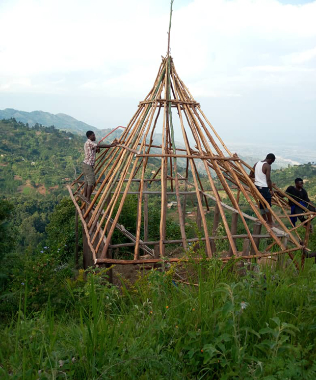 Wooden frame structure being constructed by three people on a hillside. The frame is cone-shaped, with lush greenery and distant hills in the background under a cloudy sky.