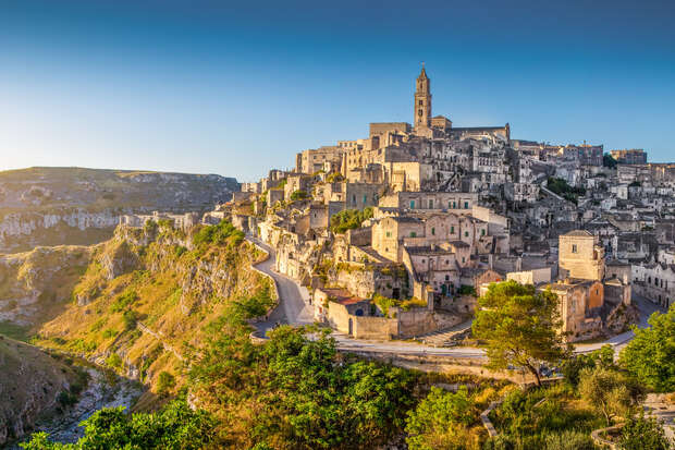Ancient stone buildings cluster on a hillside during sunset, with a prominent bell tower rising in the center; a winding path leads through lush greenery surrounding the historic town.