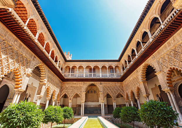 Courtyard featuring intricate Moorish architecture with detailed arches; sunlight illuminates stone carvings. Symmetrically arranged greenery surrounds a central water channel under a clear blue sky.