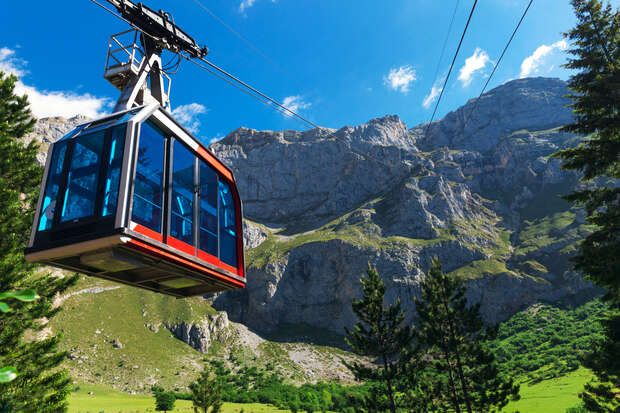 A cable car ascends with passengers, suspended above a lush green valley. In the backdrop, rugged mountains reach towards a clear blue sky, surrounded by scattered trees.