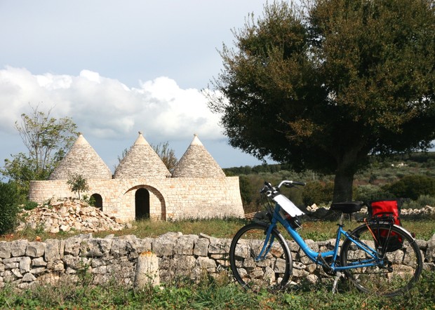 A blue bicycle rests near a stone wall in front of a trullo, a traditional dry stone hut with conical roofs, situated in a grassy rural landscape under a partly cloudy sky.