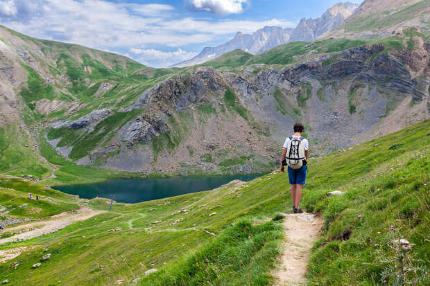 A person walks on a narrow hiking trail, surrounded by lush green hills and rocky terrain. In the background, a serene lake is nestled between peaks under a partly cloudy sky.