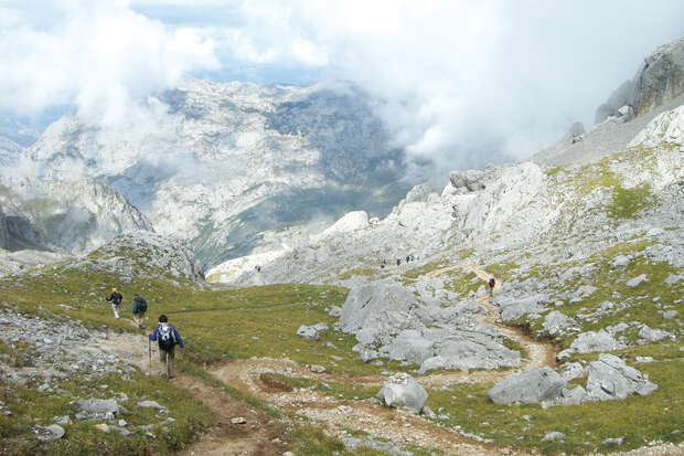 Hikers walk along a rocky trail through a mountainous landscape with green grass and scattered rocks. Clouds and distant peaks create a dramatic backdrop, enhancing the sense of wilderness.