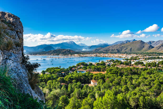 A rocky cliff overlooks a serene bay with numerous anchored boats. Lush trees and scattered buildings populate the foreground, while mountains line the horizon under a clear blue sky.
