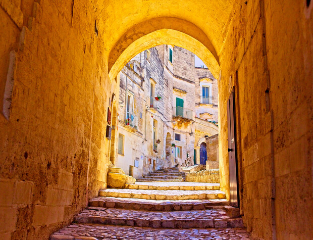 A stone archway frames a cobblestone pathway leading uphill, flanked by historical buildings with green shutters and potted plants, creating a picturesque Mediterranean village scene bathed in warm sunlight.