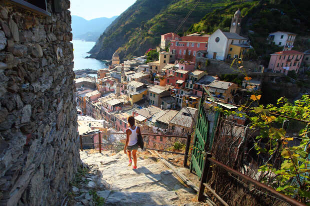 A person descends stone steps, surrounded by lush hills and colorful buildings overlooking a coastal town under clear blue skies.