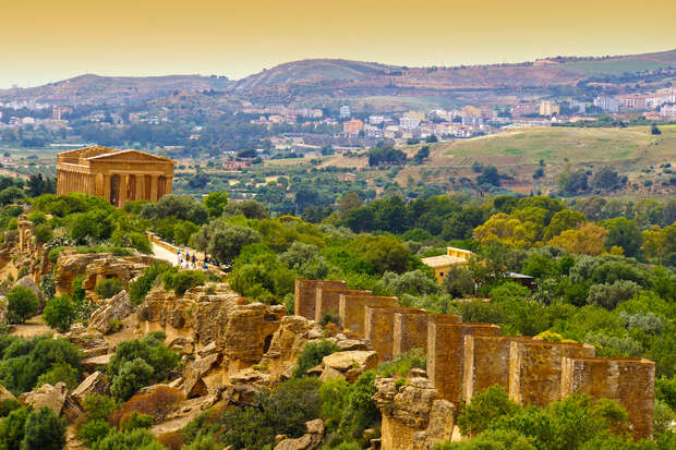 Ancient temple ruins stand among lush greenery, set against a backdrop of rolling hills and a distant town under a warm, golden sky.