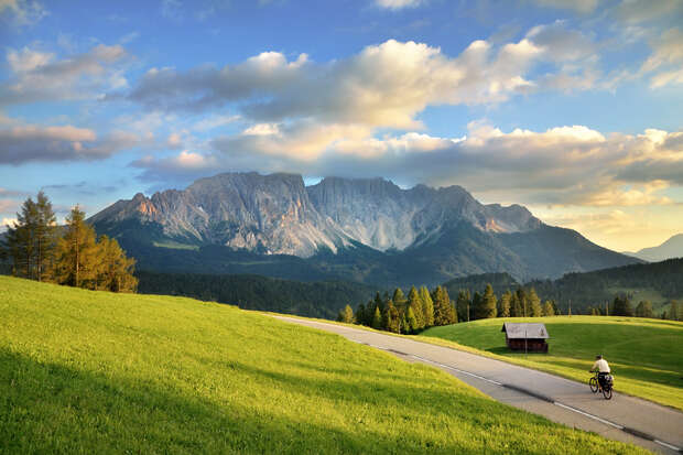 A cyclist rides along a winding road through vibrant green fields, with a small shed nearby and majestic mountains under a partly cloudy sky in the background.