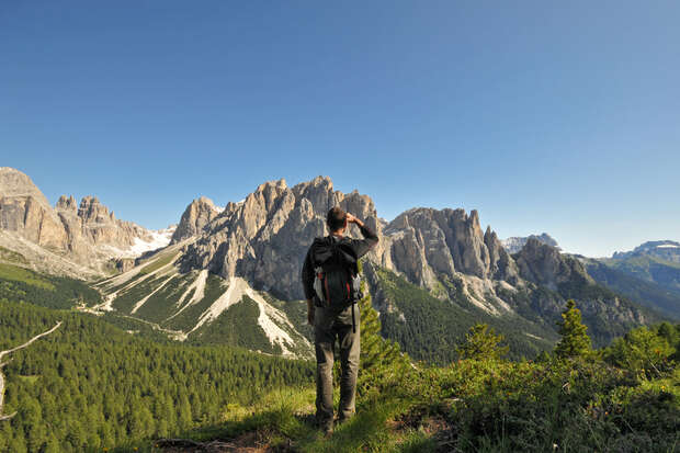 A person, wearing a backpack, faces and admires a range of rugged, sunlit mountains surrounded by lush greenery under a clear blue sky.