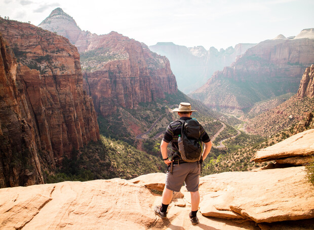 A hiker stands on a rocky ledge, overlooking a vast canyon landscape with rugged cliffs and a winding river below, under a clear sky.