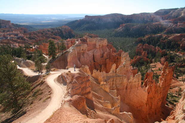 Rock formations rise dramatically, forming spires and cliffs with reddish hues. A narrow, winding path, bordered by sparse trees, leads through the natural landscape under a wide, clear sky.