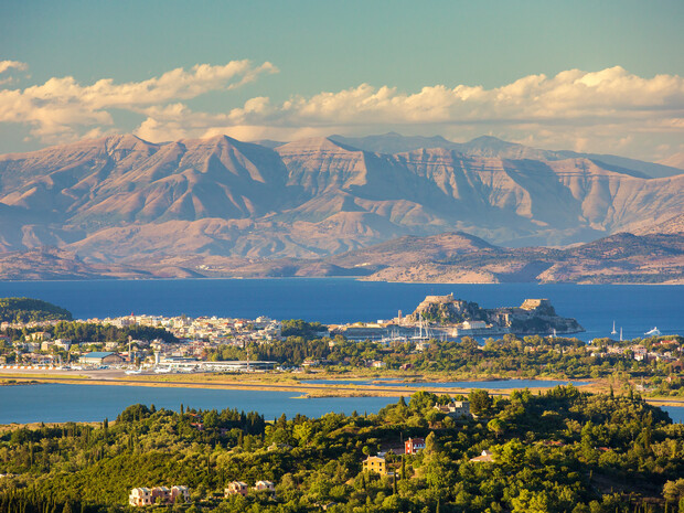 Landscape featuring a town by the sea, with a fortress on a peninsula; mountains rise in the background under a clear sky, and lush greenery covers the foreground.