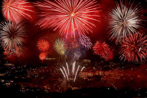 Bright fireworks burst in various colors, illuminating the night sky above a cityscape. Multiple explosions create a vivid, festive display over rooftops, celebrating an unspecified event. No text is present.