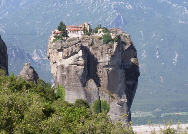 A monastery perches on top of a towering rock formation surrounded by lush greenery, with distant mountains in the background under a clear sky.