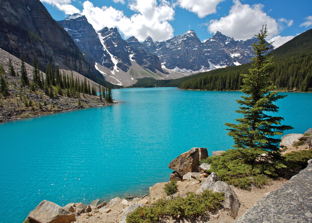 A vibrant turquoise lake sits surrounded by rugged mountains and evergreen trees under a partly cloudy sky, with rocky shores in the foreground.