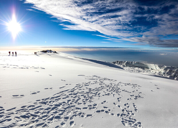 Snow-covered mountain summit with scattered footprints. Several people walk towards the peak under a bright sun in a clear blue sky. Clouds stretch across the horizon, enhancing the vast landscape.