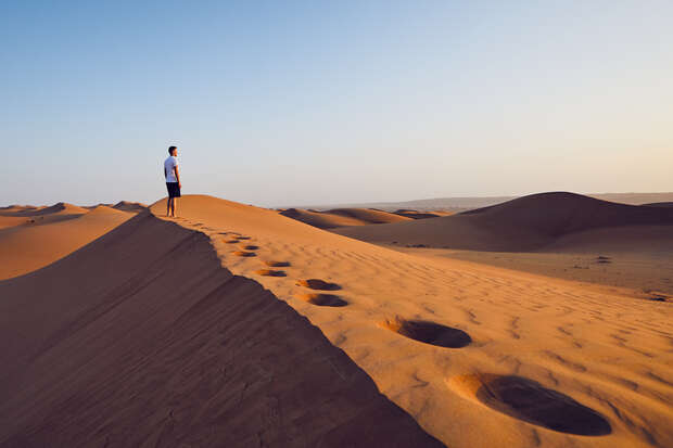 A person stands on a sunlit sand dune, surveying the vast desert landscape, with soft shadows and footprints highlighting the undulating terrain.