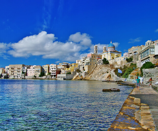 Buildings with colorful facades line a coastal cityscape, featuring a domed church on a hilltop. People walk along a stone path beside clear blue water under a bright blue sky.