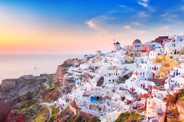 Whitewashed buildings cascade down a cliffside at sunset, with a windmill visible. The scene overlooks a calm sea, surrounded by greenery and a vibrant, colorful sky.