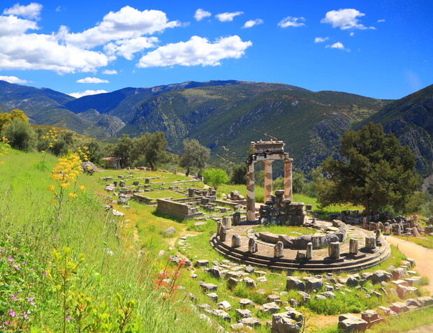 Ancient circular ruins sit in grassy terrain with wildflowers blooming. Tall columns partially stand, surrounded by scattered stone remnants. Majestic mountains and a bright blue sky with clouds form the backdrop.