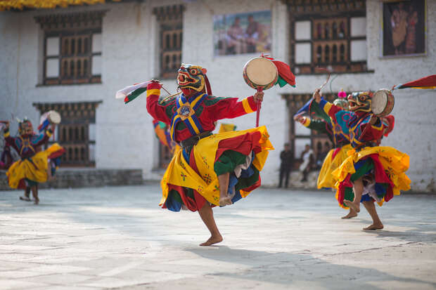 Dancers in vibrant, colorful costumes perform energetically, holding drums in a traditional courtyard with white walls and wooden windows, while a small audience watches in the background.