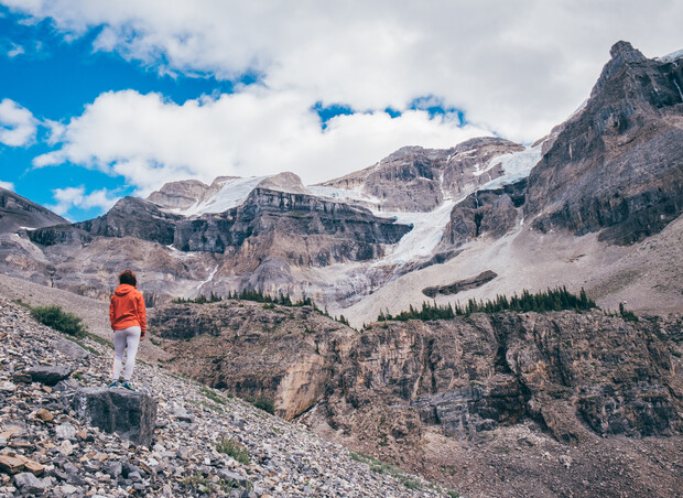 A person in an orange jacket stands on a rocky slope, gazing at rugged, snow-capped mountains under a partly cloudy sky. Sparse vegetation and layered rock formations surround the scene.