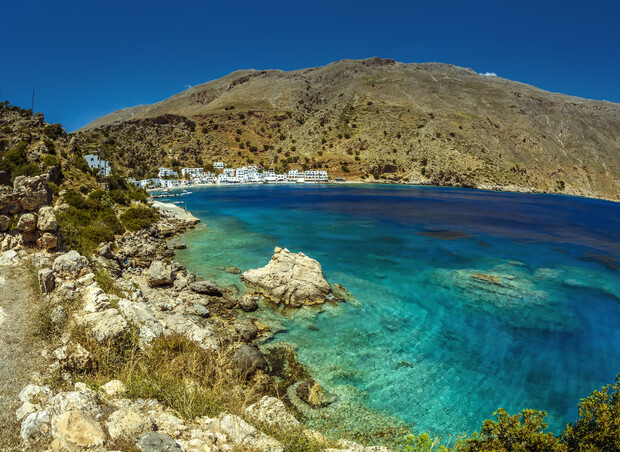 Rocky coastline borders turquoise sea, leading to a small, white-washed village nestled beneath a barren, brown hillside under a clear blue sky. No text is present.