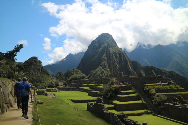 Hikers walk along a stone path beside ancient terraced structures, with a large, rugged mountain and vibrant green landscape under a partly cloudy blue sky in the background.