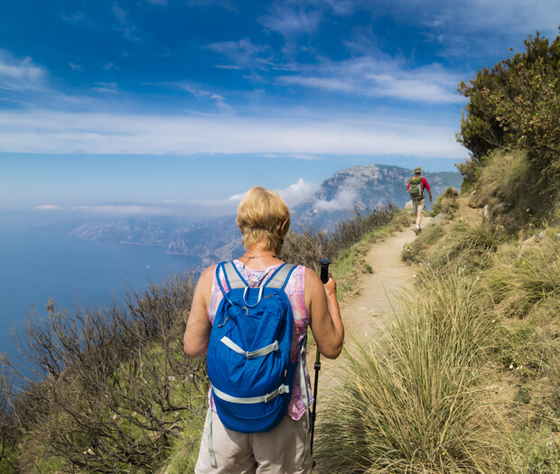 A person with a blue backpack hikes along a narrow trail, surrounded by grass and overlooking a vast ocean view on a clear, sunny day.