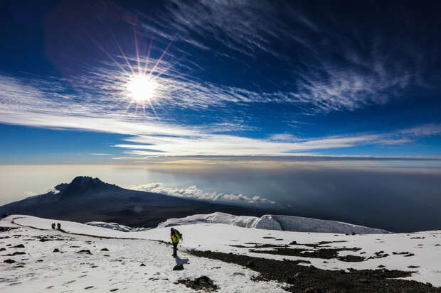 A lone hiker ascends a snow-covered mountain, silhouetted against a bright sun and expansive, cloudy sky. In the background, distant peaks rise from a misty horizon.