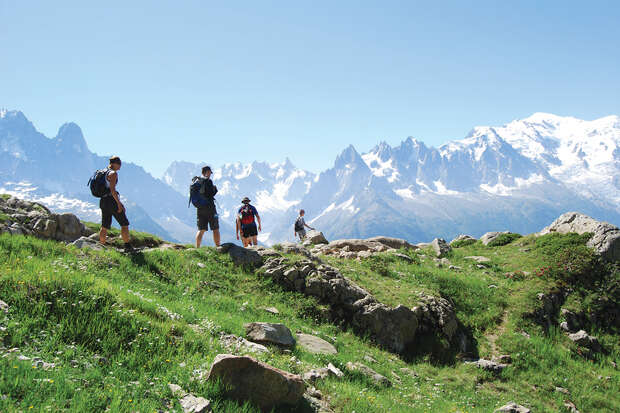 Four hikers walk along a rocky, grassy mountain trail. Snow-capped peaks are visible in the background under a clear blue sky, suggesting a scenic outdoor adventure.