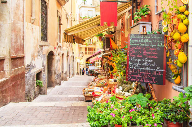 A small market stall displays vibrant fruits, vegetables, and flowers along a narrow, sunny alley. A chalkboard menu lists Italian specialties: "Bruschette ai Pomodori Pompelmi, Formaggi, Olio di Oliva..." Pedestrians walk nearby.