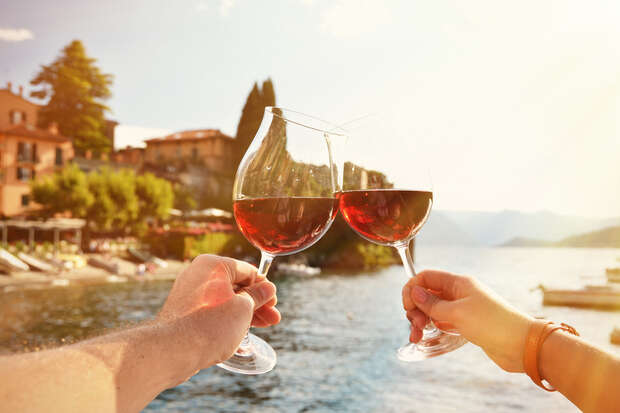 Two hands clink wine glasses filled with red wine. The setting is a picturesque lakeside scene with sunlit buildings and lush greenery in the background.