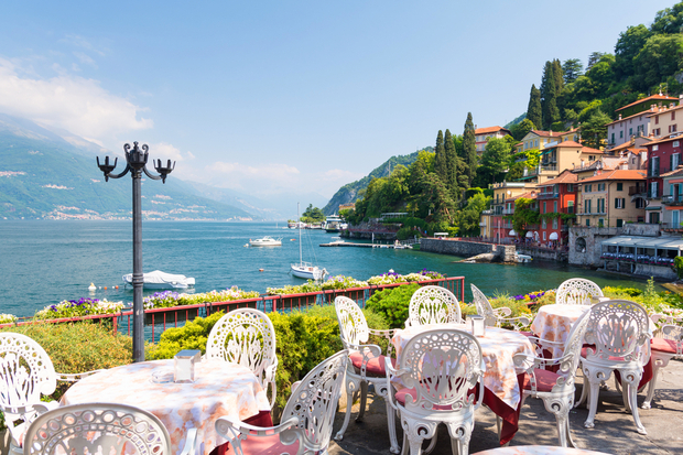 Ornate white chairs surround tables with pink tablecloths, overlooking a scenic lake. Boats float on the water near colorful hillside houses, lush greenery, and distant mountains under a blue sky.