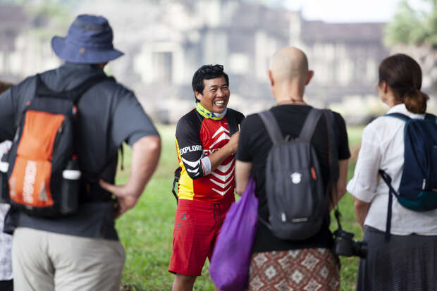 A person in colorful clothing, smiling and speaking to three other people wearing backpacks, stands in an outdoor setting with blurred ancient architecture in the background, suggesting a guided tour.