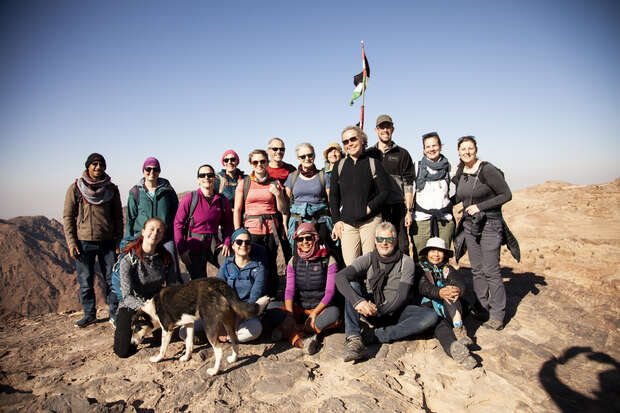 A group of people poses on a rocky summit, smiling beneath a clear blue sky. A dog stands in the foreground. A flag is visible in the background.