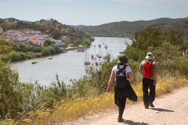 Two hikers walk along a dirt path beside a river, surrounded by lush vegetation. Hills and a whitewashed village are visible across the water under a clear sky.