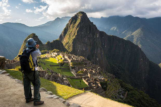 Individual photographing ancient stone ruins with a mountain backdrop. The person is standing on a path, wearing a hat and backpack. Sunlight highlights the lush landscape surrounded by misty clouds.