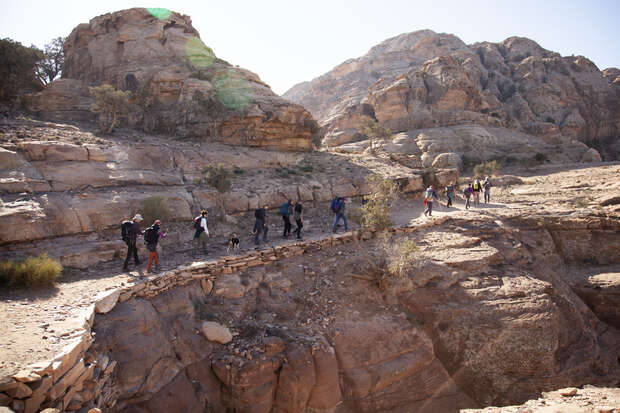 A group of hikers walks along a narrow, rocky trail with steep ledges in a mountainous, arid landscape under clear skies.