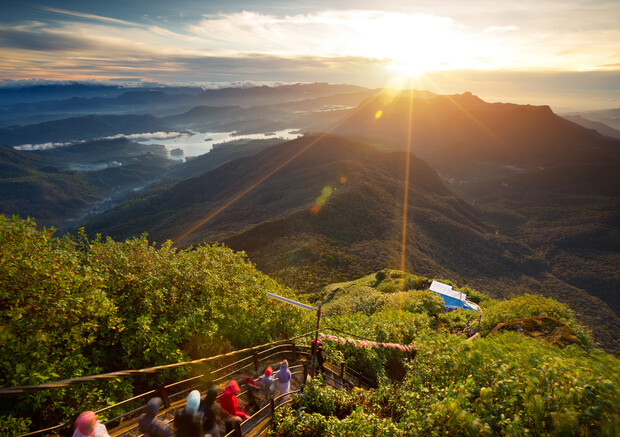 A group of hikers climbs a lush green mountain path at sunrise, with sunrays illuminating the landscape of rolling hills and distant valleys.