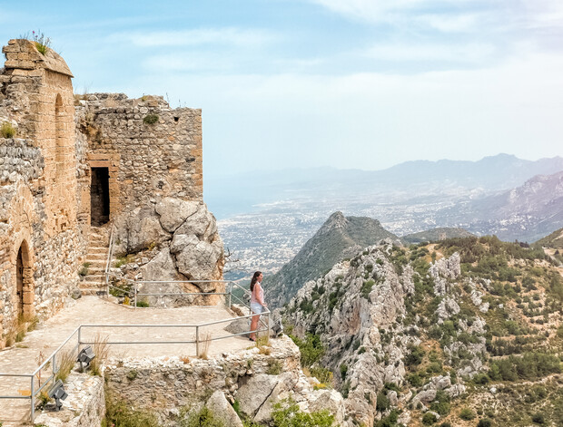A person stands on a stone terrace of an ancient castle, overlooking a vast mountain landscape and distant cityscape under a clear blue sky.
