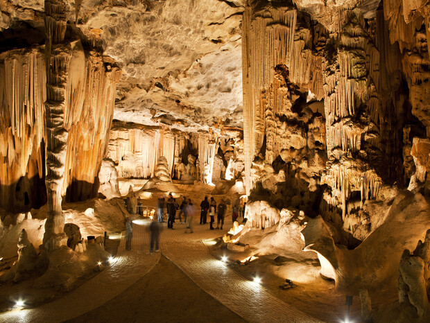 Stalactites and stalagmites tower in a dimly lit cave as a group of people explore the illuminated pathway, surrounded by intricate rock formations and textured cavern walls.