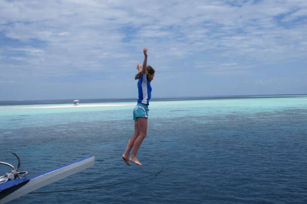 A person jumps from a boat's edge into the clear blue ocean, arms raised, surrounded by a tranquil seascape under a partly cloudy sky.
