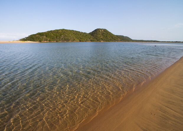 Clear water gently laps against a sandy beach, with a green, tree-covered hill rising in the background under a clear blue sky.