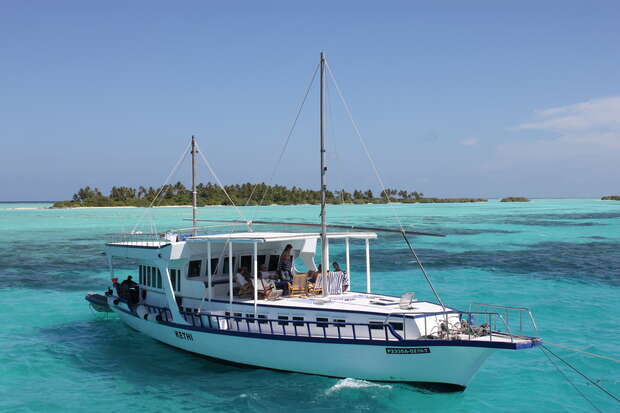 A white boat named "Bari 3" sails through turquoise ocean waters, carrying people. An island with lush greenery is visible in the background.