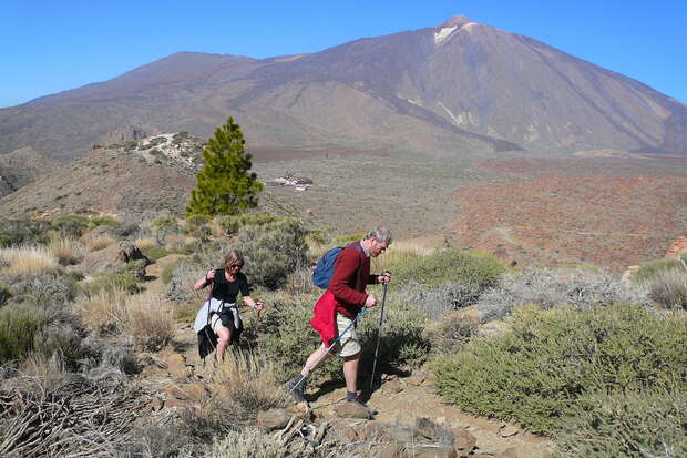 Two hikers with walking sticks trek through a rocky, shrub-filled landscape. A large mountain dominates the background under a clear blue sky.