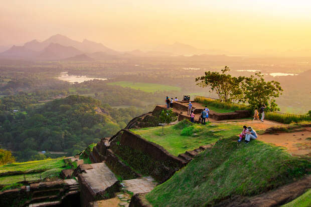 Visitors stand and sit on ancient stone ruins, overlooking a vast, lush landscape with distant mountains under a golden sky, suggesting sunset.