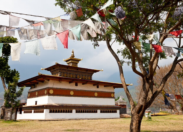 A traditional Bhutanese temple stands quietly, surrounded by colorful prayer flags hanging from trees. The structure features intricate, layered architecture against a backdrop of distant mountains and a clear blue sky.